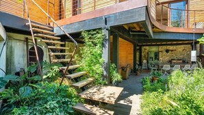Une maison moderne avec une façade en bois, un escalier menant à un balcon et un patio aménagé avec du mobilier d’extérieur.