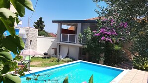 Uma piscina com superfície de água azul cristalina, rodeada por vegetação e uma casa com varanda.