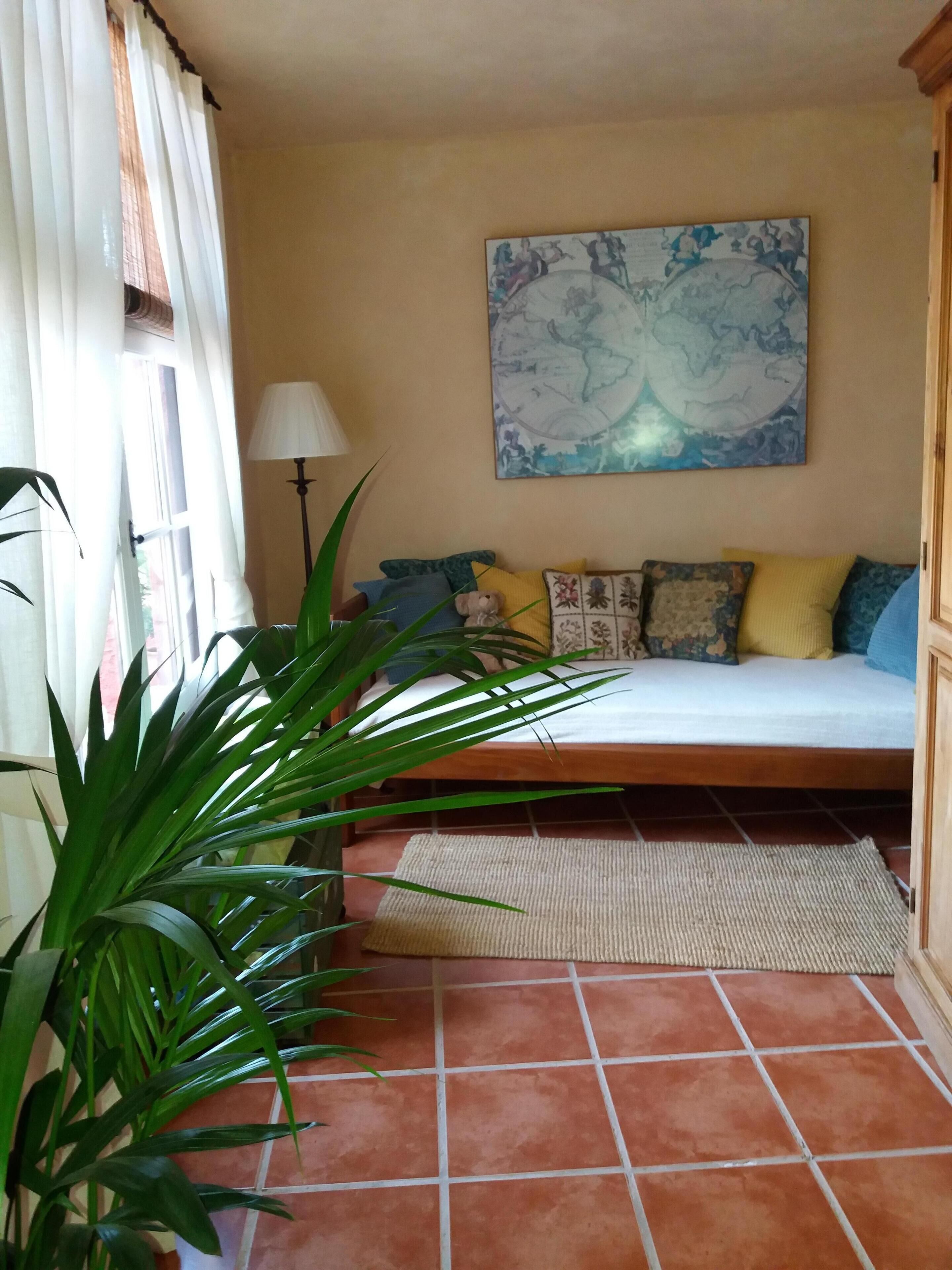 A bedroom with a bed, a large window with curtains, a floor lamp, a painting on the wall, and a potted plant.