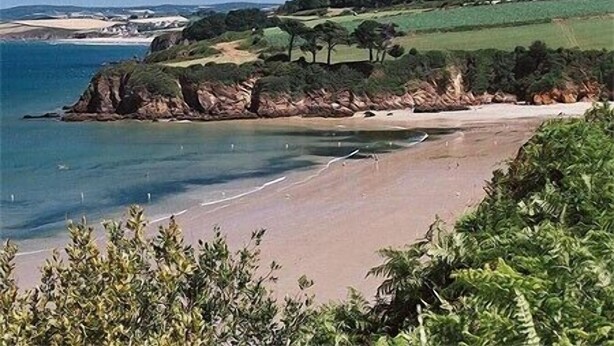 Un paysage côtier avec une plage de sable, des collines verdoyantes et un ciel bleu clair.