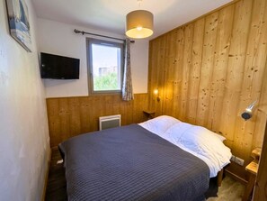 Ein Schlafzimmer mit einem Bett, einem an der Wand befestigten Fernseher, einem Fenster mit Vorhängen und einer Holzvertäfelung.