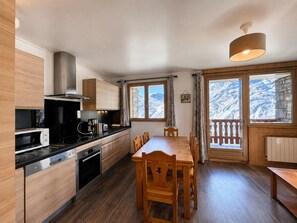 A modern kitchen with wooden cabinets, a dining table, and chairs. There is a large window with a view of snow-covered mountains. - Spacious Ski-In/Ski-Out Apartment for 6 with Balcony and Stunning Views (Les Menuires)