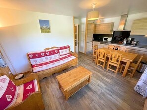 Ein gemütlicher Wohnbereich mit einer Couch, einem Holzesstisch und Stühlen. Im Hintergrund ist eine Küche mit Mikrowelle und Herd erkennbar.
