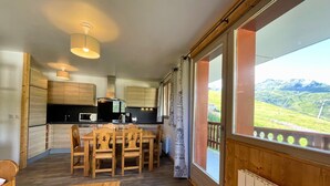 Cuina moderna amb armaris de fusta, taula de dinar amb caires i vista a un paisatge de muntanyes a través de grans finestres.