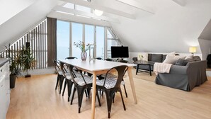 A modern loft apartment with a dining area, a living room, and a large window offering a view of the sky.