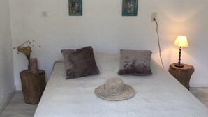 Ein Schlafzimmer mit einem großen Bett, zwei Kissen, einem gewebten Hut, einer Vase mit Blumen, einem hölzernen Nachttisch, einer Lampe und zwei an der Wand befestigten Bildern.