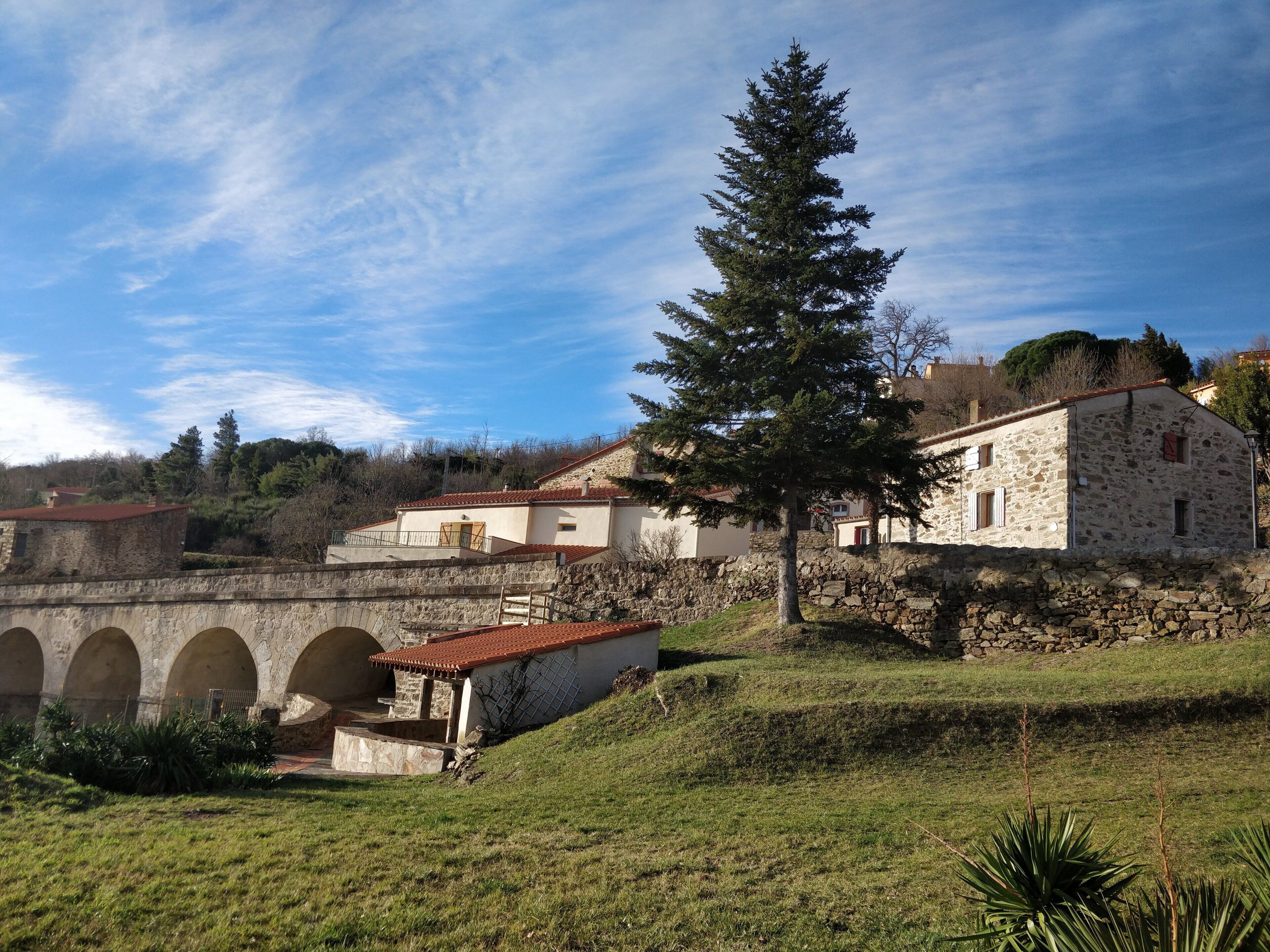 A stone bridge with arches, a stone building with a red roof, and a large evergreen tree.