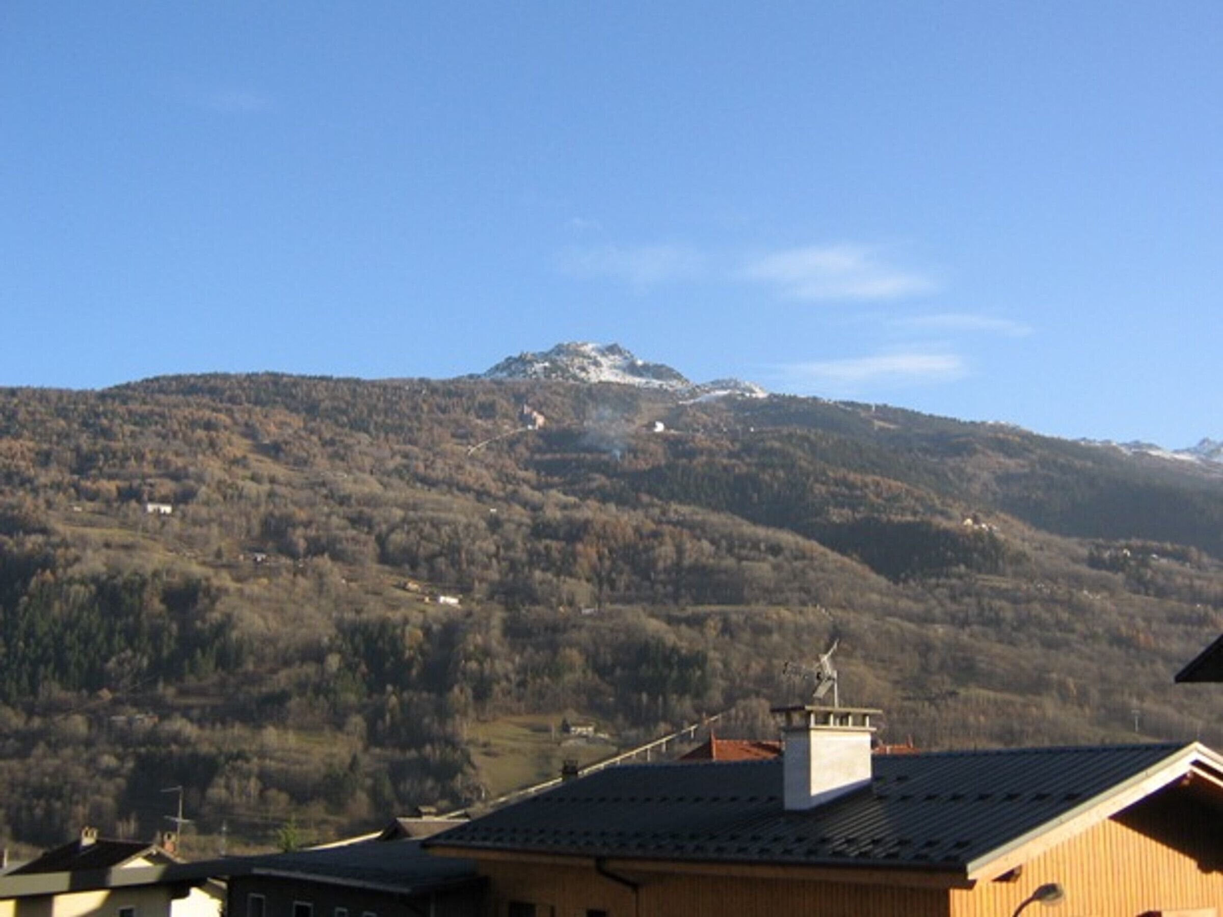 A mountain with a snow-capped peak and a clear sky. There are buildings with sloped roofs in the foreground.