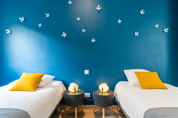 A room with two beds, each with a white bedspread and a yellow pillow. There are two black nightstands with lamps between the beds. The wall is painted blue, and there are white paper flowers on it. - LA GRANGE - CHARMING 4-PERSON GITE + 2-PERSON CABANE INSOLITE. (Birkenwald)