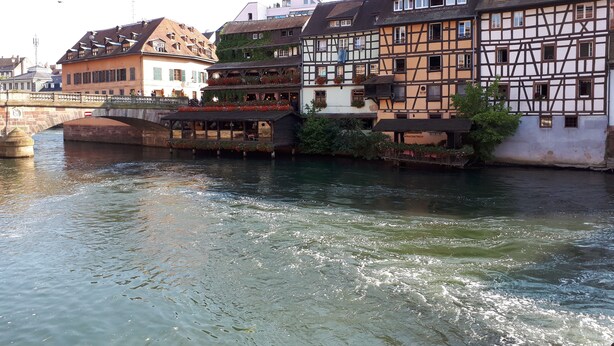 Vista a la orilla del río con edificios tradicionales de entramado de madera, un puente y un restaurante con terraza junto al agua.