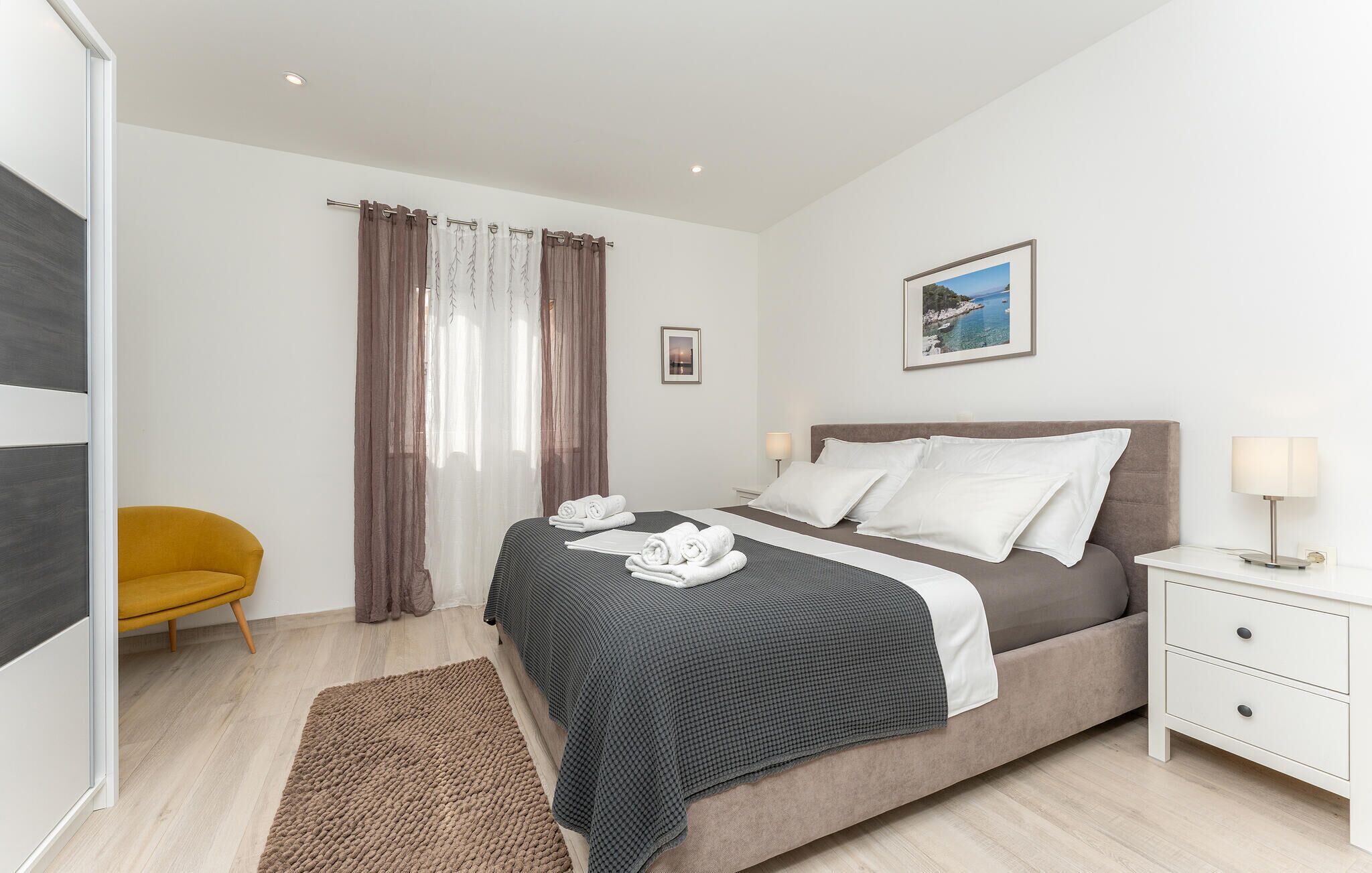 A modern bedroom with a bed, bedside tables, a nightstand, a chair, and a view of the sea.