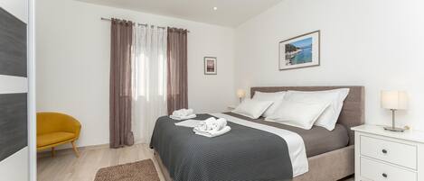 A modern bedroom with a bed, bedside tables, a nightstand, a chair, and a view of the sea.