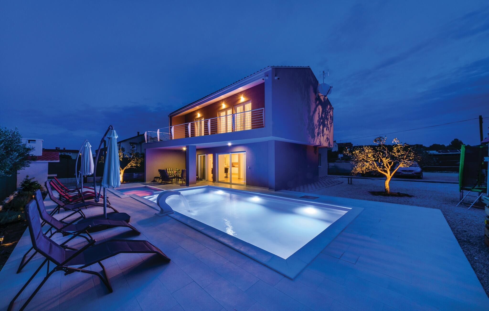 A modern two-story house with a swimming pool, outdoor seating, and a well-lit balcony.