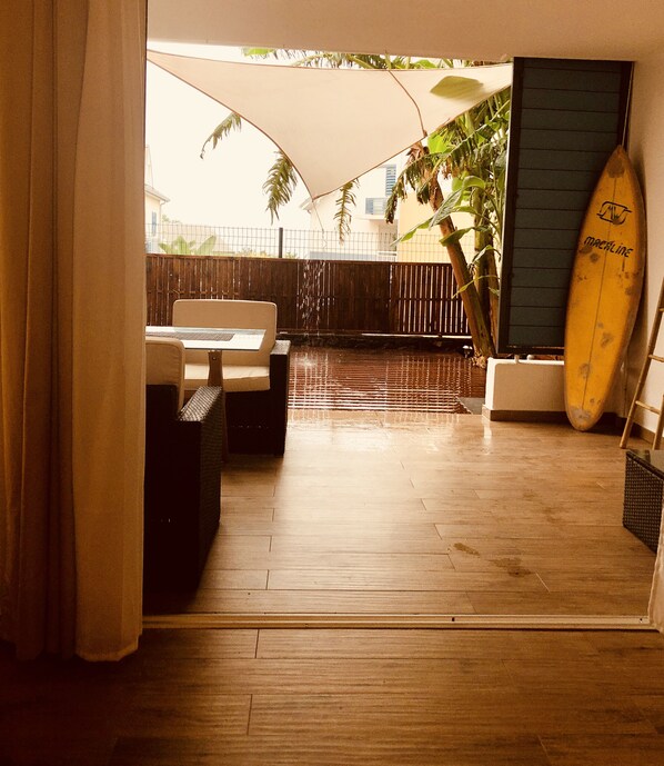 Un patio al aire libre con piso de madera, una mesa y sillas, una tabla de surf apoyada contra una pared y un paraguas grande que da sombra.