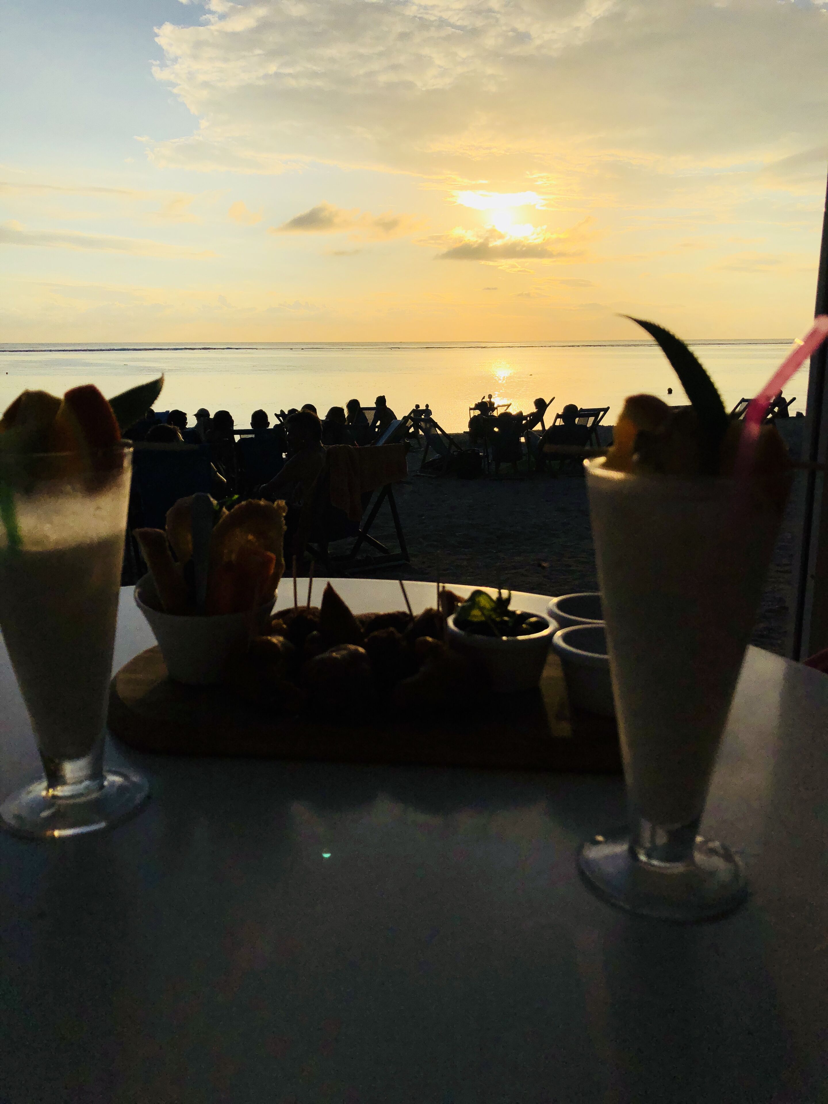 A table with two drinks and a small dish of food, overlooking a beach with people and a sunset in the background.