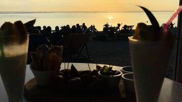 A table with two drinks and a small dish of food, overlooking a beach with people and a sunset in the background.