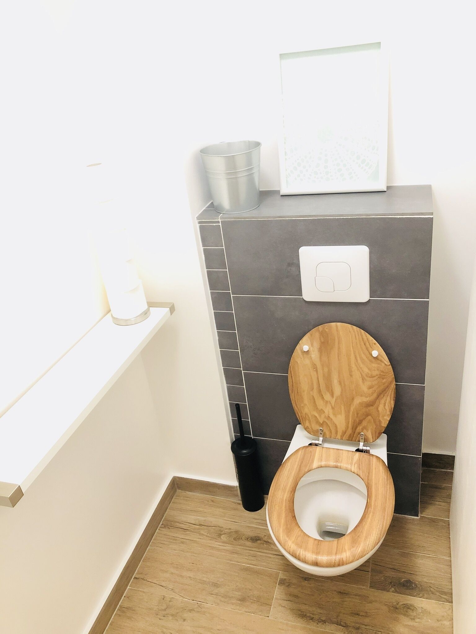 Un baño moderno con inodoro de madera, tanque de descarga empotrado en la pared y un pequeño estante con artículos de aseo.