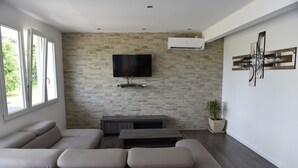 Ein modernes Wohnzimmer mit einem Ecksofa, einem Flachbildfernseher an einer Steinwand, einem kleinen Tisch und einer Zimmerpflanze.