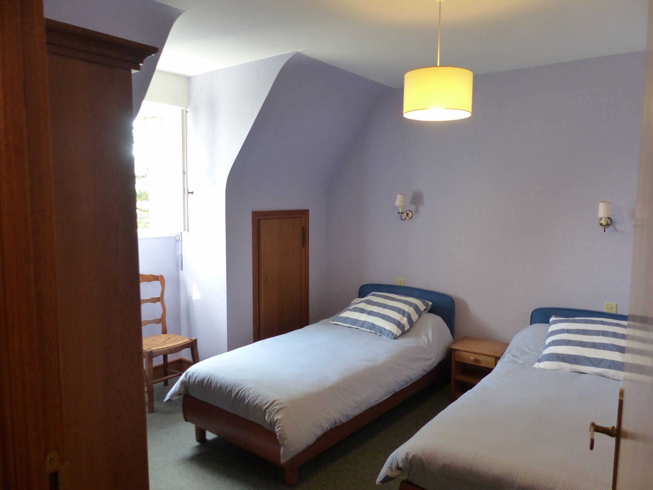 Une chambre avec deux lits, une porte en bois, une petite table et un luminaire suspendu.
