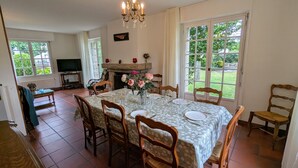 Comedor con una mesa dispuesta para seis personas, rodeada de sillas de madera. Se ve una chimenea con un jarrón de flores, y ventanas amplias que ofrecen vistas al verdor exterior.