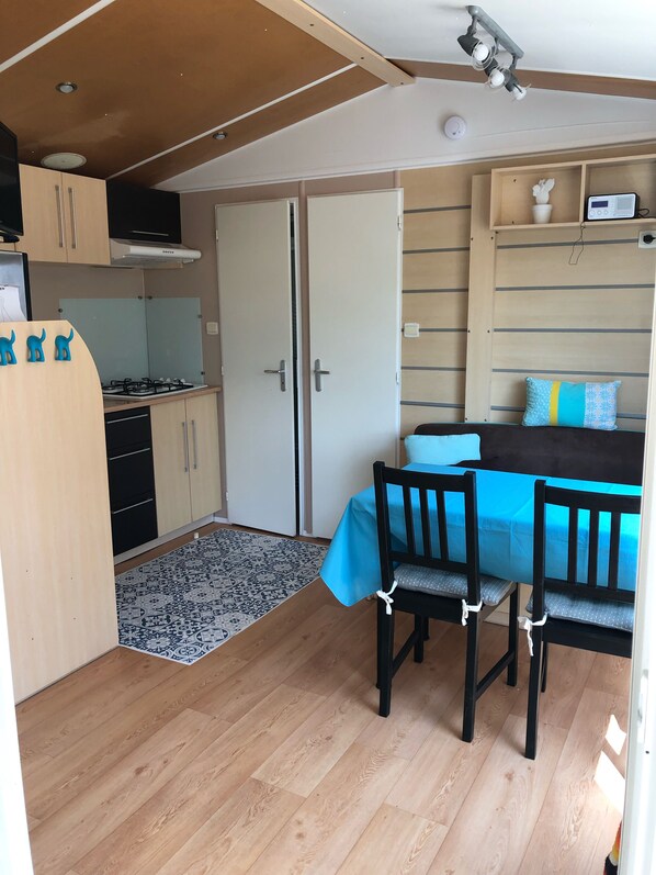 A compact living space with a kitchenette, a dining area with two chairs, a bed with a blue cover, and a small shelf with decorative items.