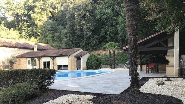 A swimming pool surrounded by a pebble border, a stone building, and a covered patio area. - Bed and Breakfast Sarlat Canada (Sarlat-la-Canéda)
