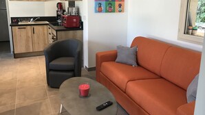 A compact living space with a kitchenette, a grey armchair, an orange sofa with a grey pillow, a small round table with a red candle, and a colorful pineapple artwork on the wall. - Bed and Breakfast Sarlat Canada (Sarlat-la-Canéda)