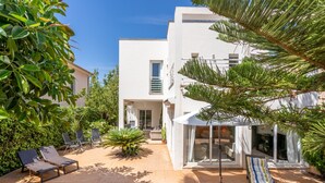 A modern building with a patio area featuring sun loungers, a small palm tree, and a glass door entrance. - Holiday Home "Villa Mar - Can Bujosa" on the Beach (Palma de Mallorca)