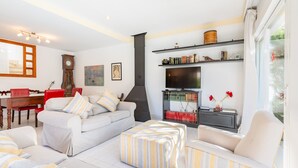 A living room with a sofa, ottoman, and armchair, a fireplace, bookshelves, and a dining area with a table and chairs. - Holiday Home "Villa Mar - Can Bujosa" on the Beach (Palma de Mallorca)