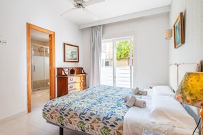 Une chambre à coucher comprenant un lit, une commode en bois, un ventilateur de plafond, une porte donnant sur un balcon et un tableau accroché au mur.