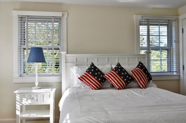 A bedroom with a white bed, a nightstand with a lamp, and two windows with blinds.