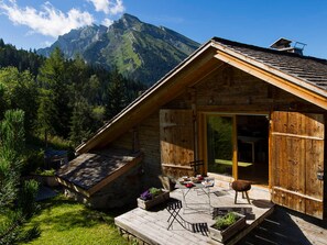 Una cabaña de madera con techo inclinado, un porche con mobiliario exterior y montañas al fondo.