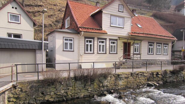 Ein zweistöckiges Haus mit rotem Ziegeldach, einer Steinsäule davor und einem Fluss, der daran fließt.
