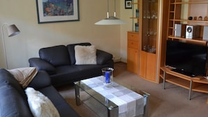 Ein Wohnzimmer mit einer schwarzen Couch, einem Glastisch, einem Fernseher auf einem Holzständer und einer Schrank mit Regalen.