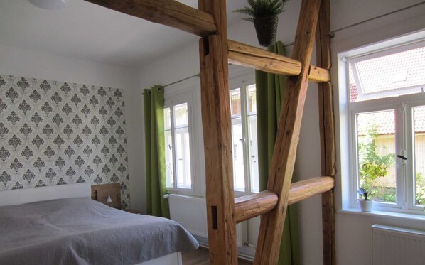 A bedroom with a bed, a wooden ladder, a window with a view of greenery, and patterned wallpaper.