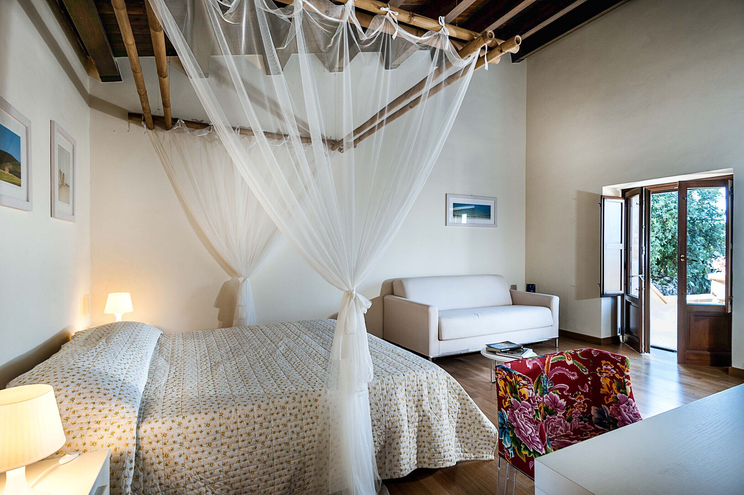 A bedroom with a bed, a white sofa, a colorful chair, a nightstand, and a window.