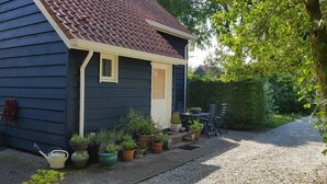 A small house with a dark blue exterior, a brown tiled roof, and a front porch with potted plants and a table with chairs.