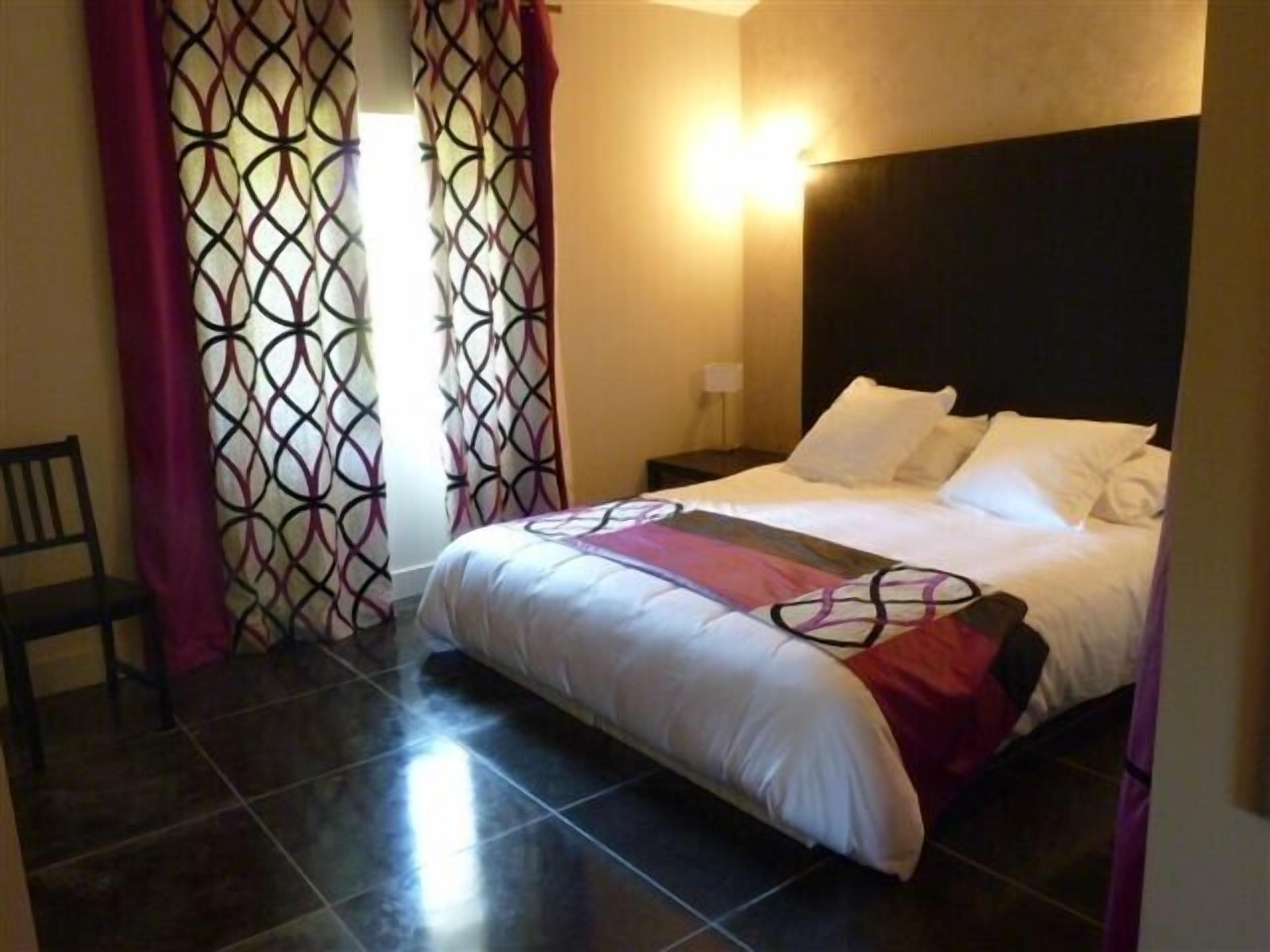 A hotel room with a bed, a nightstand, a chair, and patterned curtains.