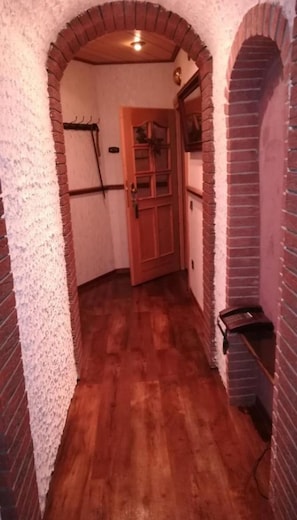 A narrow hallway with brick walls and wooden flooring, leading to a wooden door with glass panels.