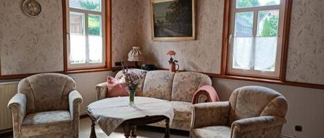 A room with a wooden ceiling, patterned wallpaper, a sofa, two armchairs, a table, and a window with a view of greenery.