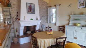 A dining area with a fireplace, wooden chairs, a table with a yellow tablecloth, and a white cabinet with decorative items. - Village house with lake view (Sainte-Croix-Du-Verdon)
