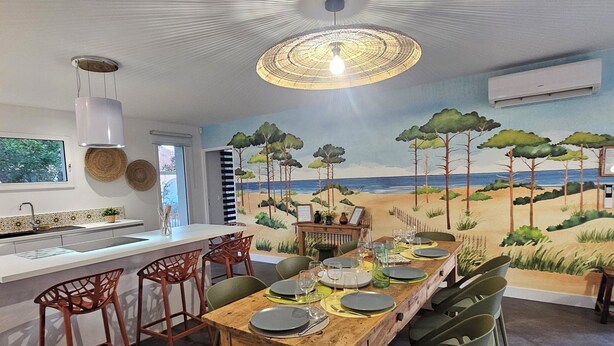 A dining area with a beach mural, a wooden table set for six, and a modern kitchen in the background. - NEW HOUSE, 700M FROM THE BEACH WITH A HEATED SWIMMING POOL AND A SPA (Labenne)