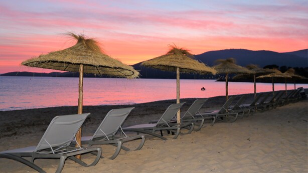 Ein Strand mit Liegestühlen und Reetdachschirmen mit Blick auf ein Gewässer bei Sonnenuntergang.