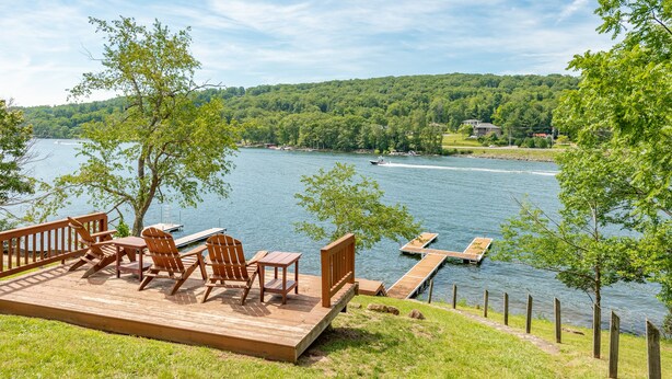 A wooden deck with outdoor furniture overlooking a lake, surrounded by greenery and a dock. - Spacious Lake Home w/ Dock, Hot Tub & Views (McHenry)