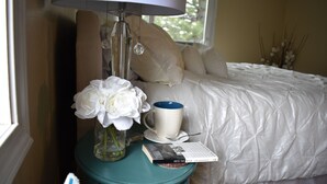 Una cama bien hecha con una colcha blanca y almohadas, una mesita de noche color turquesa con una lámpara, un jarrón con flores blancas, una taza y un libro.