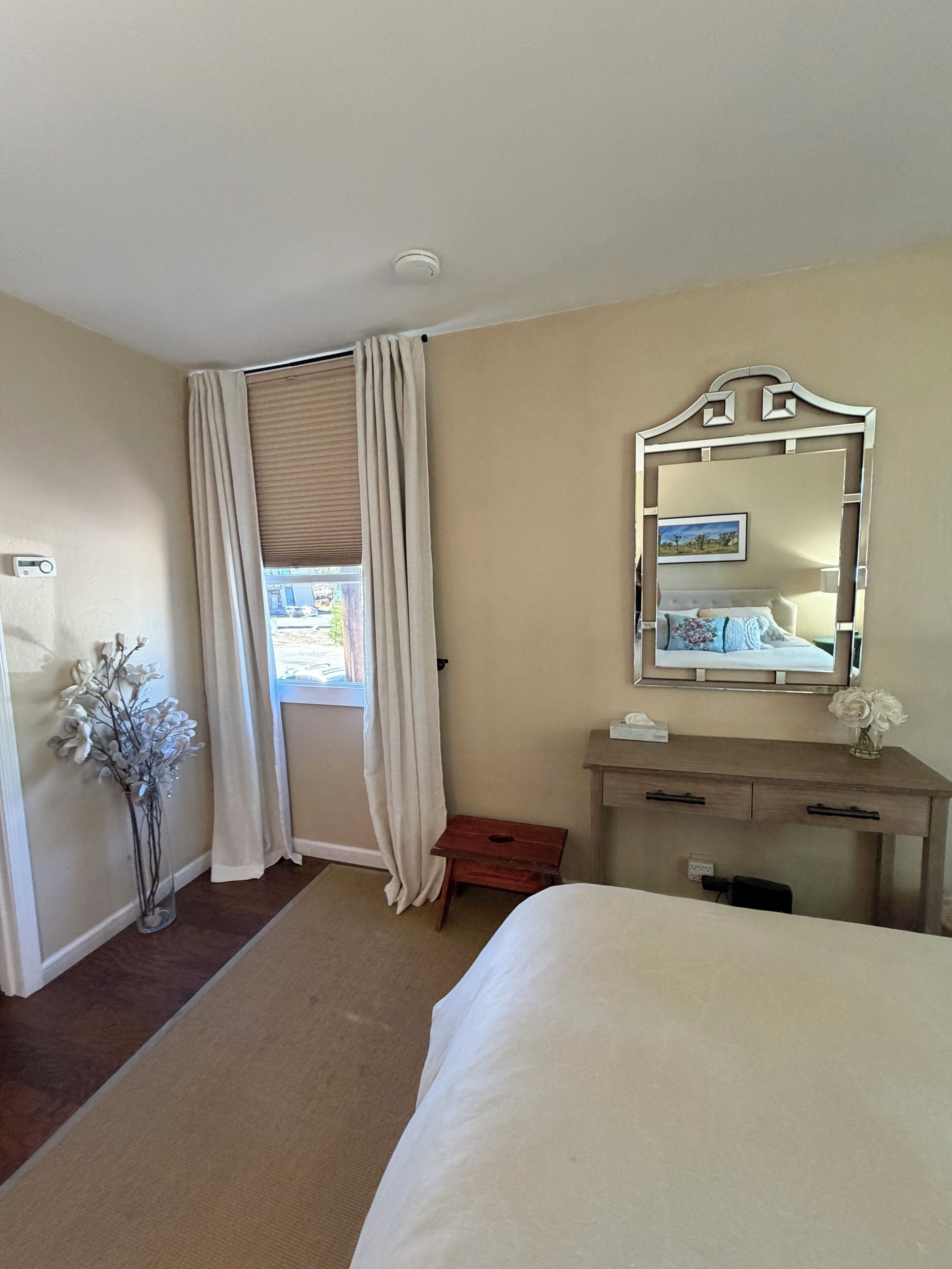 A hotel room with a bed, a bedside table, a mirror, a window with curtains, and a vase with flowers.