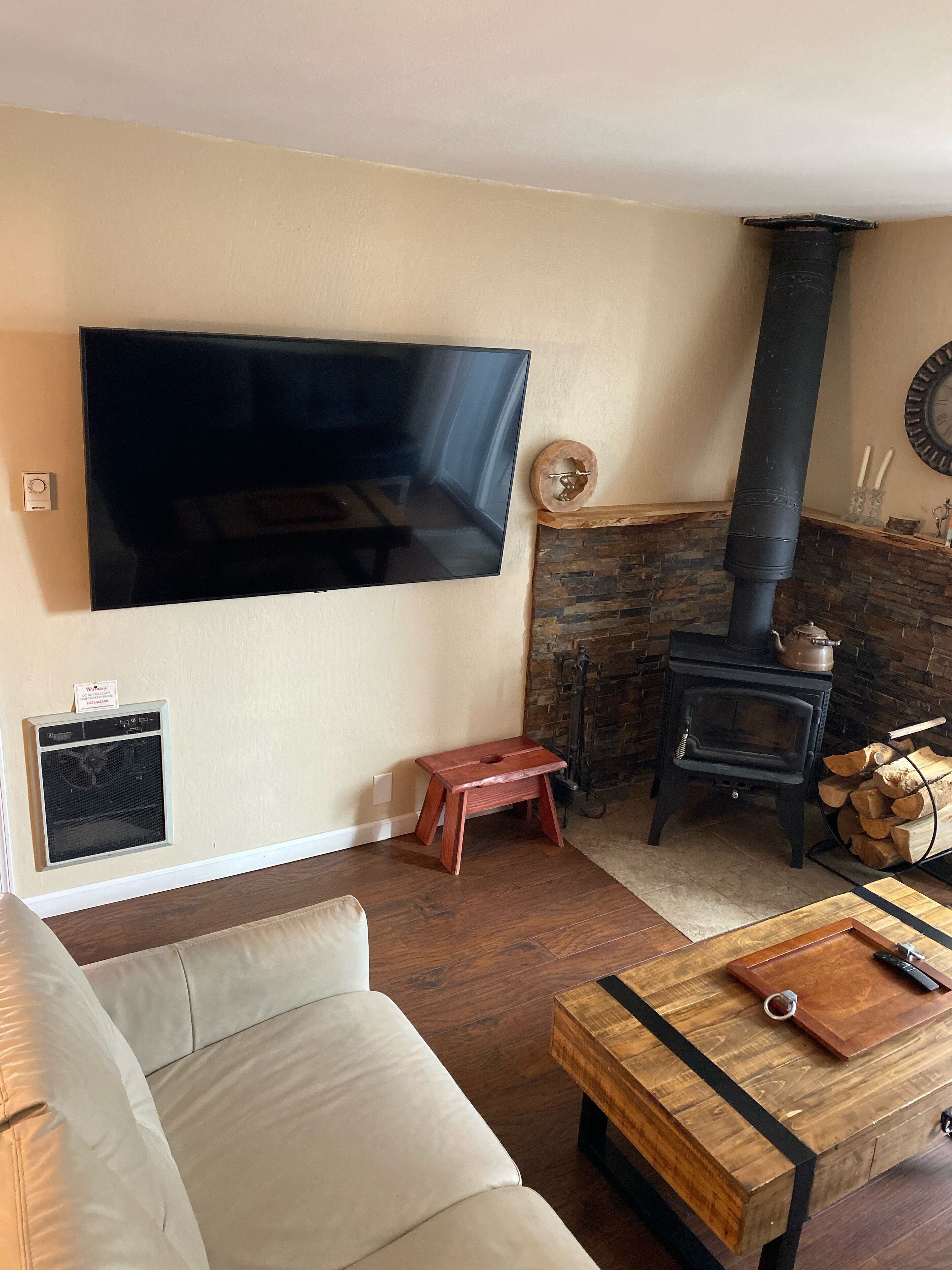 Ein Wohnzimmer mit Flachbildfernseher an der Wand, einem Steinkamin, einem schwarzen Kaminofen, einem Holztisch mit Messer und Schneidebrett sowie einem beigefarbenen Sofa.