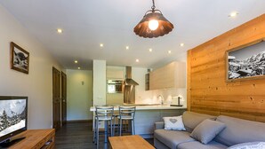 Una sala de estar moderna con piso de madera, un sofá, una mesa de centro y un televisor sobre un mueble de madera. Cuenta con una zona de cocina con estufa, lavaplatos y armarios.