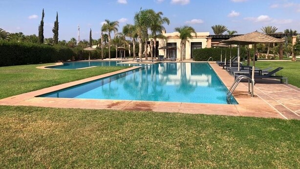 A rectangular swimming pool with a tiled deck, surrounded by a well-maintained lawn and palm trees. - VILLA "LOUISA" 4P-PRIVATE POOL GROUND FLOOR PARKING COMPLEX DYARSHEMSI  (Oulad Teima)