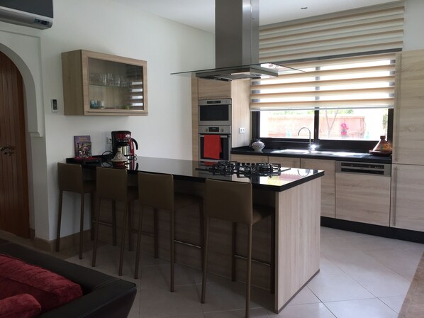 A modern kitchen with a central island, bar stools, a coffee machine, and a view through the window. - VILLA "LOUISA" 4P-PRIVATE POOL GROUND FLOOR PARKING COMPLEX DYARSHEMSI (Oulad Teima)